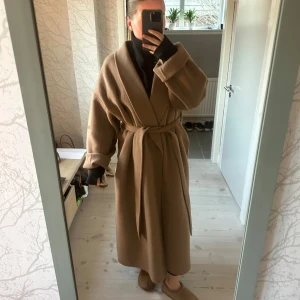 Ullkappa med knytband - Säljer en stilren beige/camel kappa med knytband i midjan. Den har en omlott-design och långa ärmar, perfekt för kyligare dagar. Passar bra för både höst och vinter. Ifrån Hm, sparsamt använd. Storlek L men går att dra åt så det blir en smalare passform. Är själv en M vanligtvis 