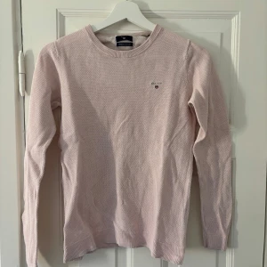 Ljusrosa tröja från GANT - Säljer en stilren ljusrosa tröja från GANT i premium bomull. Tröjan har en rund halsringning och ett diskret broderat GANT-emblem på bröstet. Perfekt för en avslappnad och snygg look.