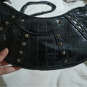 Svart axelväska med nitar - Snygg svart axelväska i syntet med krokodilpräglat mönster och coola nitar. Perfekt för att ge din outfit en edgy touch. Väskan har en justerbar axelrem och stängs med dragkedja.