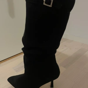 Svarta stövlar med klack och dragkedja - Eleganta svarta stövlar i mocka med spetsig tå och klack. De har en dekorativ spänne upptill och en praktisk dragkedja på sidan för enkel på- och avtagning.