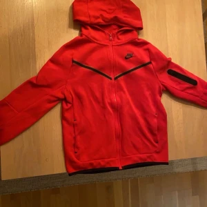 Röd nike tech kofta  - Säljer min röda nike tech kofta då den är för liten för mig. Den är i helt okej skick men ett litet hål på baksidan som man inte märker av. Stl xl i barn,159-170cm 