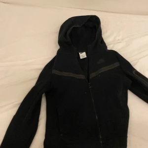 Svart huvtröja från Nike - Säljer en svart huvtröja från Nike med dragkedja och huva. Perfekt för en sportig look eller avslappnade dagar. Tröjan har en enkel design med diskreta detaljer.