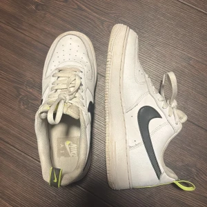 Vita Nike Air Force sneakers - Säljer ett par vita Nike Air Force sneakers i bra skick. De har en klassisk design med svart swoosh och gula detaljer på hälen och tungan. Perfekta för vardagsbruk och ger en stilren look. Skorna har snörning och är gjorda i skinn.