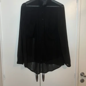 Svart blus från Gina Tricot - Snygg svart blus från Gina Tricot i transparent material. Blusen har långa ärmar och knappar framtill. Perfekt för en stilren look.