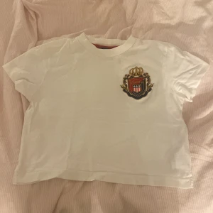 Vit croppad t-shirt från Gant - Säljer en vit croppad t-shirt från Gant med ett broderat emblem på bröstet. Perfekt för en avslappnad stil. Passar åldern 11-12 år. ❄️🪽