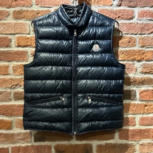 Navy dunväst från Moncler - Säljer en snygg navy dunväst från Moncler med dragkedja och två praktiska fickor framtill. Västen är i herrstorlek 2 vilket motsvarar S/M. Västen har en quiltad design och Moncler-logga på bröstet. Perfekt för kyligare dagar! 