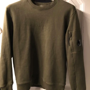 Olivgrön tröja från C.P. Company - Säljer en snygg olivgrön tröja från C.P. Company. Den har en cool detalj med en ficka på ärmen och en rund logotyp. Perfekt för höst och vår, och den är oandvänd . Passar både till vardags och lite mer avslappnade tillfällen. Den är 100% äkta, taggar och kvitto finns,
