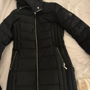 Svart dunjacka med dragkedja - Snygg svart dunjacka med långa ärmar och praktiska dragkedjor på sidorna. Perfekt för kalla dagar med sin quiltade design och höga krage.