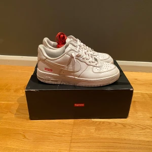 Nike Air Force 1 Low Supreme - Säljer ett par vita Nike Air Force 1 Low i samarbete med Supreme. Skorna är i bra skick och har den klassiska Air Force 1-designen med Supreme-loggan på sidan. De kommer med extra röda skosnören för en cool kontrast. Skorna är sparsamt använda. 