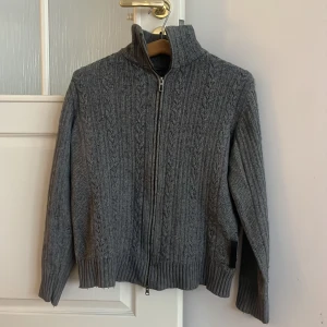 Grå stickad kofta från Marc O'Polo - Säljer en stilren grå stickad kofta från Marc O'Polo med dragkedja och hög krage. Perfekt för kyliga dagar med sitt klassiska kabelstickade mönster och ribbade kanter. Priset kan diskuteras vid snabb affär