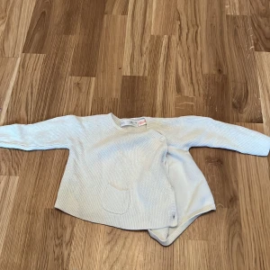 Vit stickad babytröja från Zara - Söt vit stickad babytröja från Zara med långärmad design och en liten ficka framtill. Perfekt för att hålla din lilla varm och bekväm under kyligare dagar. Tröjan har knappar på sidan för enkel på- och avklädning.