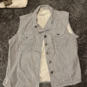 Rutig väst med knappar - Säljer en stilren rutig väst i svart och vit med knappar framtill. Västen har två bröstfickor med knappar och en klassisk krage. Perfekt för en avslappnad look eller som ett lager under hösten.