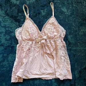 Rosa spetslinne med rosett - Säljer ett romantiskt rosa linne med spetsdetaljer och en söt rosett framtill. Linnet har justerbara axelband och är perfekt för en mysig hemmakväll eller som ett lager under en tröja. Det är i mycket bra skick och ger en feminin touch till din garderob.