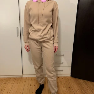 Beige hoodie och byxor i mysigt material - Säljer en supermysig set  i mjukt material. Perfekt för chilliga dagar hemma eller en avslappnad dag ute. Den har en klassisk passform med långa ärmar och en praktisk huva med dragsko. 