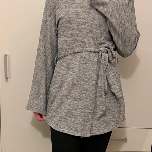 Grå kofta med knytband - Säljer en stilren grå kofta i mjukt material. Koftan har långa ärmar och ett knytband i midjan för en justerbar passform. Perfekt för höst och vår när du vill ha något lätt men ändå snyggt. Passar både till vardags och lite finare tillfällen.