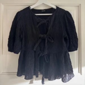 Svart blus med knytning - Säljer en svart blus med knytning framtill. Den har trekvartsärmar och ett fint, krusigt mönster i tyget. Perfekt för både vardag och festliga tillfällen. Passar bra till jeans eller kjol! 