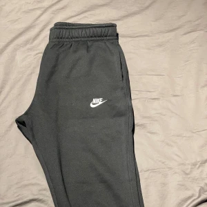 Svarta mjukisbyxor från Nike - Säljer ett par svarta mjukisbyxor från Nike i mycket bra skick. De har en elastisk midja och en praktisk bakficka med knapp. Perfekta för en avslappnad stil eller träning. Nike-loggan är snyggt placerad på benet. Sköna och stilrena byxor för alla tillfällen! Pris kan diskuteras