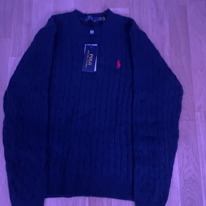 Svart kabelstickad tröja från Polo Ralph Lauren - Snygg svart kabelstickad tröja från Polo Ralph Lauren med det ikoniska röda logotypbroderiet på bröstet. Tröjan har långa ärmar och en klassisk rund halsringning. Perfekt för en stilren look. Mycket snygg tröja. Helt ny aldrig använd! Scan fungerar perfekt! Pris kan diskuteras vid snabb affär!
