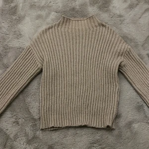 Beige ribbad stickad tröja - Säljer en mysig beige stickad tröja med ribbad design. Tröjan har långa ärmar och en hög krage som ger extra värme. Perfekt för kyliga dagar.