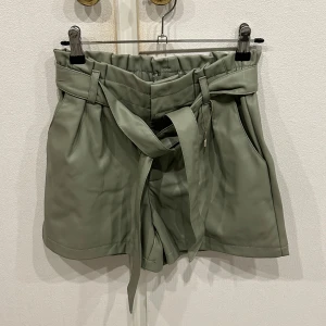 Olivgröna shorts med knytband - Snygga olivgröna shorts med hög midja och knytband i midjan. Perfekta för en stilren look. Shortsen har fickor på sidorna och en bekväm passform. Helt nya aldrig använda!