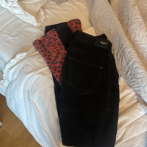 Svarta jeans med röd leopardmönster från Pepe Jeans - Snygga svarta jeans från Pepe Jeans med en unik röd leopardmönstrad detalj längs benen. Perfekta för att ge din outfit en cool twist. De har en klassisk femficksdesign och knappgylf.