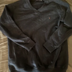 Mörkblå tröja från Tommy Hilfiger - Säljer en klassisk mörkblå tröja från Tommy Hilfiger med diskret logga på bröstet. Tröjan har långa ärmar och ribbade muddar vid ärmslut och nederkant. Perfekt för en stilren look.