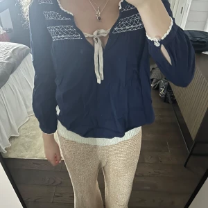 Mörkblå blus med knytband - Charmig mörkblå blus med vita broderier och knytband framtill. Blusen har långa ärmar med dekorativa detaljer vid ärmsluten. Från oss Molly