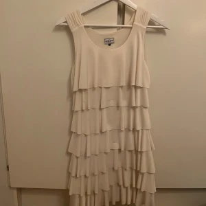 Vit klänning med volanger - Elegant vit klänning med flera lager av volanger. Den är ärmlös och har en rund halsringning, vilket ger en stilren look. Perfekt för sommarens festligheter.