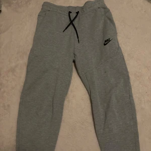 Grå mjukisbyxor från Nike - Säljer ett par grå mjukisbyxor från Nike med svart snörning i midjan och en dragkedja på sidan. Perfekta för en avslappnad stil.