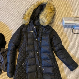 Rockandblue Svart dunjacka med päls - Snygg svart dunjacka med quiltade detaljer och en mysig pälsfodrad huva. Jackan har dragkedja och knappar framtill samt praktiska fickor med dragkedja. Perfekt för kyliga dagar.