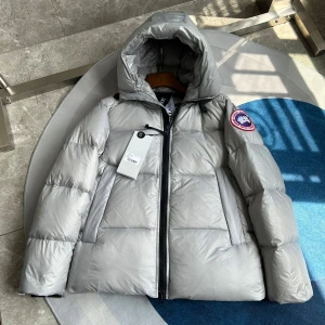 Grå dunjacka från Canada Goose - Säljer en stilren grå dunjacka från Canada Goose med huva och dragkedja. Jackan har en ikonisk logga på ärmen och är perfekt för kalla dagar. Den är fodrad med äkta päls och har praktiska fickor.