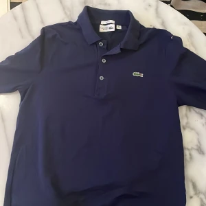 Mörkblå pikétröja från Lacoste - Säljer en stilren mörkblå pikétröja från Lacoste Sport. Tröjan har en klassisk krage med tre knappar och det ikoniska gröna krokodilemblem på bröstet. Perfekt för en sportig och avslappnad look.