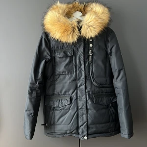 Svart parkajacka med fuskpäls från Hollies - Snygg svart parkajacka från Hollies med stor, fluffig avtagbar fuskpäls på huvan. Jackan har flera praktiska fickor och stängs med dragkedja och knappar. Perfekt för kalla dagar. Små defekter finns (se bilder).