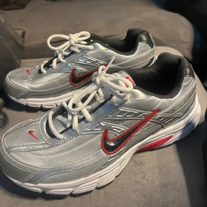 Nike silverfärgade sneakers - Snygga silverfärgade Nike sneakers med röda och svarta detaljer. Skorna har en klassisk snörning och en bekväm sula, perfekt för en sportig look.