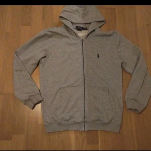 Grå hoodie från Ralph Lauren - Säljer en stilren grå hoodie från Ralph Lauren med dragkedja och det klassiska logotypbroderiet på bröstet. Perfekt för en avslappnad look. Den har långa ärmar och en bekväm passform.