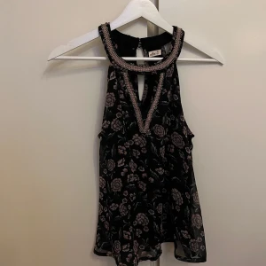 Svart blommig topp från Hollister - Snygg svart ärmlös topp från Hollister med blommigt mönster i rosa och beige. Toppen har dekorativa pärldetaljer runt halsringningen och en liten öppning i ryggen. Perfekt för en stilfull look.