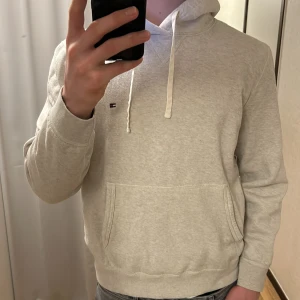 Grå hoodie från Tommy Hilfiger - Säljer en stilren grå hoodie från Tommy Hilfiger med klassisk logga på bröstet. Tröjan har en justerbar huva med vita snören och en praktisk magficka. Perfekt för en avslappnad look.