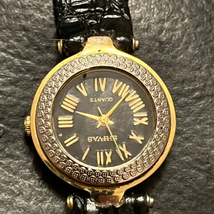 Shivas Guldfärgad klocka med svart armband - Elegant guldfärgad klocka med svart armband i krokodilmönstrat skinn. Urtavlan har romerska siffror och en glittrig kant. Perfekt för att ge en lyxig touch till din stil.