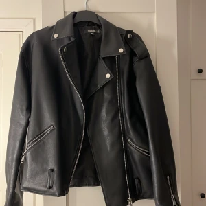 Svart skinnjacka - Snygg svart skinnjacka med klassisk bikerstil. Jackan har dragkedja framtill och på fickorna samt stiliga knappar på kragen. Perfekt för en cool look.