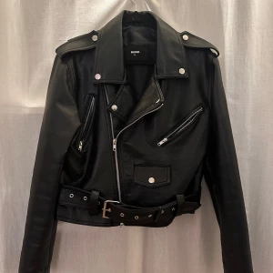 Svart skinnjacka från BikBok - Snygg svart skinnjacka från BikBok med dragkedjor och bälte. Klassisk bikerstil med silverfärgade detaljer och axelklaffar. Perfekt för en edgy look. KÖPAREN STÅR FÖR FRAKT 📦 