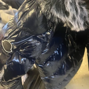 Svart dunjacka från Moncler - Snygg och glansig svart dunjacka från Moncler med pälskantad huva. Perfekt för kalla dagar med sin quiltade design och dragkedja framtill. En stilren jacka som håller dig varm och trendig. Priser kan diskuteras och det finns många storlekar av denna jacka