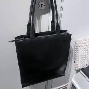 Svart läder väska med dragkedja - Snygg och stilren svart axelväska i skinn med praktisk dragkedja upptill. Väskan har två handtag och en rymlig insida med fack för dator och två andra lite mindre fack. 