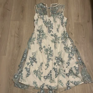 Blommig klänning från Phase Eight - Elegant klänning från Phase Eight i en ljus färg med vackra blommiga broderier i blått och grönt. Klänningen har en ärmlös design och ett medföljande bälte som ger en fin siluett. Perfekt för speciella tillfällen.