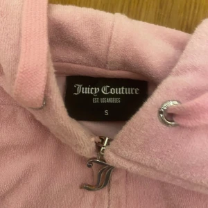 Rosa hoodie från Juicy Couture - Säljer en söt rosa hoodie från Juicy Couture med dragkedja och huva. Den har långa ärmar och en praktisk ficka framtill. Perfekt för en avslappnad stil. Handuks matrila