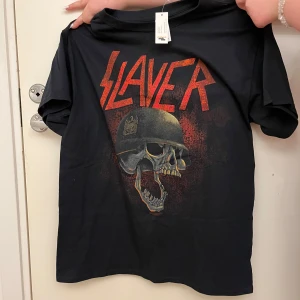 Svart Slayer t-shirt från Gildan - Cool svart t-shirt från Gildan med Slayer-tryck i rött och en dödskalle med hjälm. Perfekt för fans av bandet! Tillverkad i bomull för en bekväm passform.