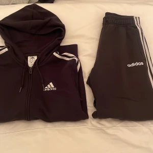 Svart hoodie och byxor från Adidas - Säljer en svart hoodie och matchande byxor från Adidas. Hoodien har dragkedja och klassiska vita ränder på ärmarna. Byxorna har elastisk midja och vita ränder längs sidorna. Perfekt för en sportig look!