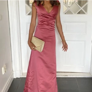 Rosa balklänning med v-ringning - Elegant rosa balklänning med djup v-ringning och breda axelband. Klänningen har en figurnära passform och är tillverkad i ett glansigt material som ger en lyxig känsla. Perfekt för en bal. Köpt här på plick. Lånade bilder från den jag köpte från. Lite använd. 32/xxs