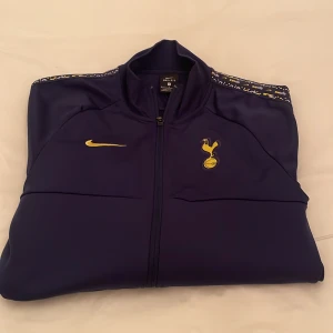 Blå träningsjacka från Nike - Snygg blå träningsjacka från Nike med Tottenham Hotspur-logga och gul Nike-swoosh. Jackan har dragkedja och långa ärmar med mönstrad detalj på axlarna. Perfekt för fotbollsfans! Mycket bekväm att träna i.