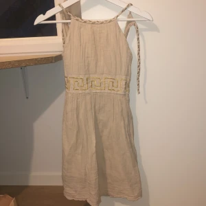 Beige klänning med guldmönster - Charmig beige klänning med ett unikt guldmönster runt midjan. Klänningen har smala axelband och en flätad detalj som ger en bohemisk känsla. Perfekt för varma sommardagar.Köpte klänningen i en marknad i Grekland då jag inte vet helt säkert vad det är för storlek men skulle säga xs