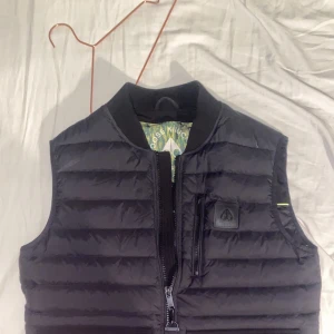 Svart dunväst från Moose Knuckles - Köpt på Johnells i Linköping. Säljer en stilren svart dunväst från Moose Knuckles. Västen har en praktisk dragkedja framtill och en bröstficka med dragkedja. Perfekt för kyligare dagar och ger en modern look.